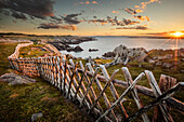 A rocky Atlantic shoreline with a wooden fence in a criss-cross pattern lines the shore as the golden sun rises in the distance; Newfoundland, Canada