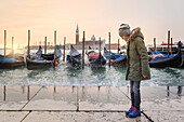 Little girl plays among the sea splashes near the gondolas docked on the Grand Canal, Venice, Veneto, Italy