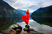 Blonde girl in a red dress moved by the wind standing on a rock in front of a lake, Eidfjord, Hordaland, Norway