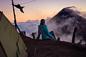 Rear view of young female hiker sitting outside tent and looking at view at summit Acatenango Volcano at sunset, Guatemala