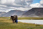 rider getting his horse ready near a lake surrounded by mountains, tavan bogd massif, altai, bayan-olgii province, mongolia