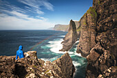 hiker contemplating the cliffs of vagar island in front of an agitated sea and a slight rainbow, sorvagsvatn, leitisvatn, vagar, faroe islands, denmark