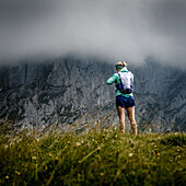 Young woman with running clothes is standing on a grassy hill in front of rock walls of Wilder Kaiser, stormy weather,Scheffau, Tyrol, Austria