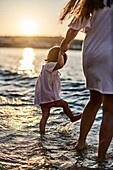 Mother holding hand of daughter at beach, standing in sea water, sunset.