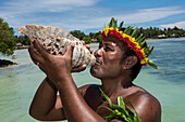 Ein Mann mit Blumen- und Blattkrone bläst in eine große Muschel, um die Ankunft von Besuchern eines Expeditionskreuzfahrtschiffes anzukündigen, Butaritari Atoll, Gilbert-Inseln, Kiribati, Südpazifik