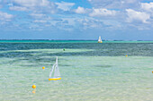 A yellow toy sailboat navigates shallow waters in the foreground while an almost identical full-sized boat sails in deeper waters in the distance, Rarotonga, Cook Islands, South Pacific