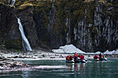 Passengers of an expedition cruise ship in Zodiac dinghy rafts boats approach a beach with a prominent waterfall, penguins, Antarctic fur seals (Arctocephalus gazella) and southern elephant seals (Mirounga leonina), Hercules Bay, South Georgia Island, Ant