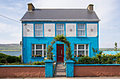 Ein Rosenbogen lenkt den Blick auf die rote Haustür eines blau-weiß gestrichenen Häuschen am Slea Head Drive, gesehen von einer Wanderung entlang dem Weitwanderweg Dingle Way, Dingle, Dingle Halbinsel, County Kerry, Irland, Europa
