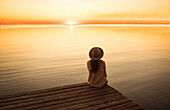 Junge Frau mit Hut sitzt an einem Steg und genießt den Sonnenuntergang