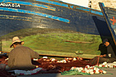 Fishermen are sitting in the harbor of Essaouira on the ground and fixing nets in front of a colored boat, Essaouira, Morocco