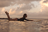 Frau auf Surfbrett liegend beim Surfen in Ozean in der Dämmerung, Kuta, Bali, Indonesien