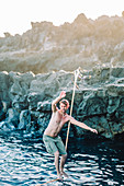 Young shirtless man balancing across tightrope over coastal water,†Tenerife, Canary Islands, Spain