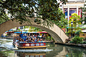 A crowd of people ride in a water taxi along the San Antonio River walk on a sunny late summer day.