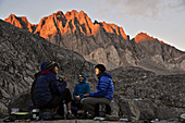 Backpackers have dinner with headlamps sitting at camp on the Barrett Lakes in Palisade Basin on a two-week trek of the Sierra High Route in Kings Canyon National Park in California. The 200-mile route roughly parallels the popular John Muir Trail through the Sierra Nevada Range of California from Kings Canyon National Park to Yosemite National Park. Sunset lights up the North Palisade in the background. 
