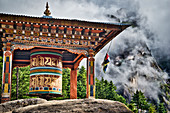Prayer wheel on a rock in the forest on the ascent to the monastery Taktshang (in the background) or Taktsang or Tigernest, a Buddhist monastery in the Parotal, Bhutan, Himalayas, Asia