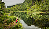 Hiker crouching at still lake in remote mountains, Faja Grande, Flores, Portugal