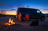 Frau am Lagerfeuer und Van an den Sanddünen von Kelso im Mojave Nationalpark bei Sonnenuntergang\n