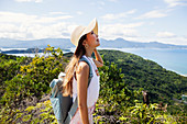 Japanese woman wearing hat and carrying backpack standing on a cliff, ocean in the background.