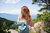 Rear view of woman carrying backpack standing on a rock on cliff, ocean in the background.