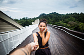 Lächelnde junge Frau steht auf einer Brücke, verdeckt ihr Gesicht und hält Männerhand
