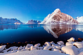Schneebedeckte Gipfel spiegeln sich im gefrorenen Meer in sternenklarer Winternacht, Reine Bay, Nordland, Lofoten, Arktis, Norwegen, Skandinavien, Europa