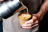 Person mit tätowiertem Finger bereitet Café Latte (Milch aus Metallkrug ins Glas gießen)