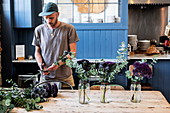 Man wearing baseball cap standing at a table, cutting thistles for flower arrangements in large glass jars.