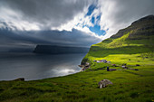 Stürmischer Himmel über den steilen Fjordhängen, Sonnenstrahl beleuchtet einen Betrieb au einem grünen Hang, Kalsoy, Faroe Islands, Dänemark