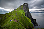 HIKER ON A STEEP PATH NEAR THE KALLUR LIGHTHOUSE IN FRONT OF THE ABRUPT SEA CLIFFS, KALSOY, FAROE ISLANDS, DENMARK