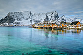 Orange-farbene Fischerhäuser vor verschneiten Bergen, Sakrisoya, Lofoten, Nordland, Norwegen