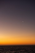 Ein Sichelmond geht auf, als der letzte warme Schein der Sonne am Horizont schwindet, nahe Puerto Chacabuco, Aysén, Patagonien, Chile, Südamerika