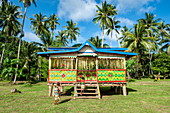 Two people enter a Selzen house with bright colors and patterns in a grassy landscape surrounded by palm trees, Garove Island, Vitu Islands, West New Britain Province, Papua New Guinea, South Pacific