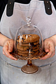 Close up of person wearing apron holding domed glass cake stand with stack of chocolate chip cookies.
