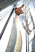 A man in white shorts with bare chest climbing up the rigging of a sailing boat.