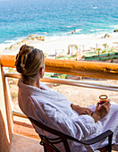 Adult woman sitting with a drink on a hotel balcony overlooking a blue ocean and white sand beach