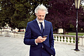 A man wearing a suit and tie standing in a walkway looking at a mobile phone.