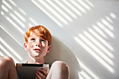 Boy with red hair sitting on floor in sunny room, holding digital tablet.