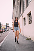 Portrait of young woman with dark brown hair, wearing black vest and denim shorts, walking down street.