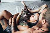 Bearded tattooed man with long brunette hair and woman with long brown hair cuddling with fluffy grey cat on a sofa.