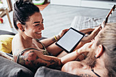 Bearded tattooed man with long brunette hair and woman with long brown hair sitting on a sofa, looking at digital tablet.