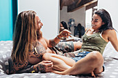 Bearded tattooed man with long brunette hair and woman with long brown hair sitting on a bed, smiling at each other.