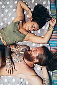 Tätowierter Mann mit langen brünetten Haaren und Bart und Frau mit langen braunen Haaren auf einem Bett liegend lächeln sich an