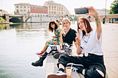 Drei junge Frauen sitzen an einem Flussufer und machen Selfie