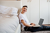 Young woman with shaved head sitting in bedroom, using laptop, smiling at camera.