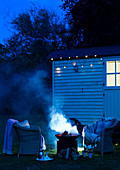 Nachtaufnahme Feuer in einer Feuerschale vor einem alten Holzbungalow, arrangiert mit Korbstühlen und Decken, Bray, Großbritannien