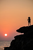 Silhouette of young woman wearing conical hat and looking out to sea from rock ledge next to Dinh Cao Shrine at sunset, Duong Dong, Phu Quoc Island, Kien Giang, Vietnam, Asia