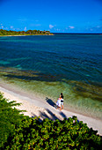 A couple walking along a sandy beach in a tropical setting. Shot from above with a view of the ocean and the bay.\nAntigua, West Indies.