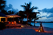 An evening shot of a bonfire set on the beach in front of a beach-terracelit with tiki-lights