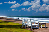 Two, white deck chairs set along a beach with a surfer and his board in the far background.\nShot in Seminyak, Bali, Indonesia