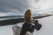 Rückansicht einer Frau, die einen Neoprenanzug trägt und ein Surfbrett hält, das auf einem verschneiten Strand steht und auf Meer schaut.
